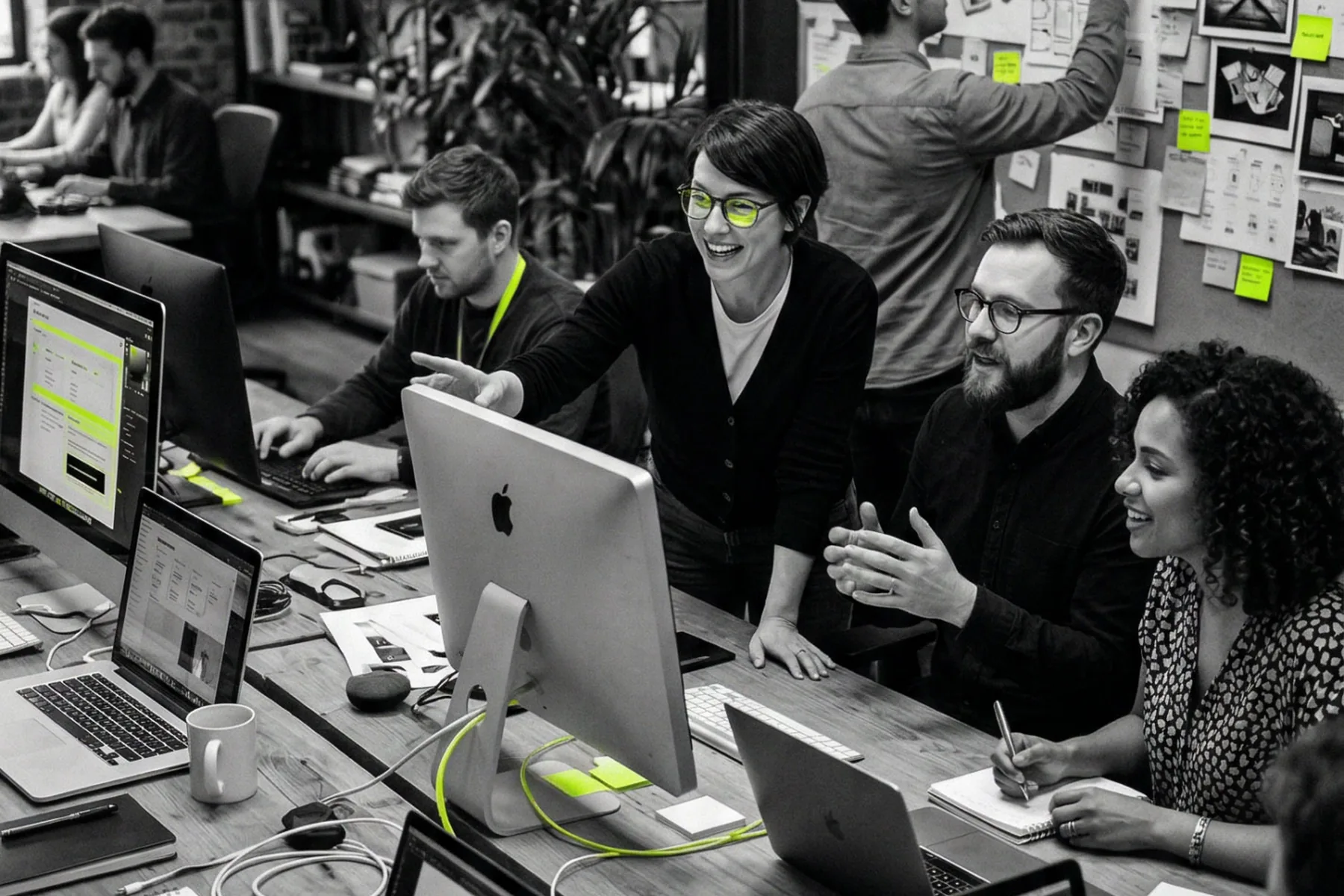 Designers gathered around a shared studio desk reviewing layouts, a wall of printed references and notes behind them.
